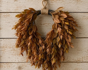 May include: A heart-shaped wreath constructed from numerous small, pointed, brown and golden-toned leaves. The wreath is suspended by a thick, natural-colored jute rope that is knotted at the top and wrapped securely around the center of the frame.