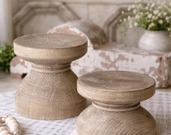 May include: Two wooden pedestals with a natural finish. Each pedestal has a round top and a flared base, creating a unique hourglass shape. The pedestals are placed on sheet music, suggesting a rustic or farmhouse aesthetic.