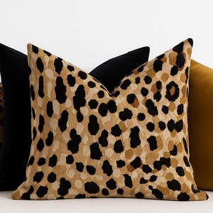 May include: A collection of decorative throw pillows in various colors and patterns. The central pillow features a tan and black leopard print. Flanking it are a black pillow and a mustard-yellow pillow. The pillows are arranged on a white surface.