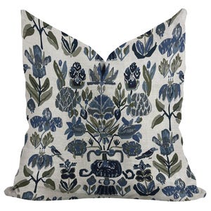 以下が含まれることがあります： クリーム色の背景に、青、緑、グレーの色合いの花柄が施された正方形の装飾用クッション。 デザインは、様式化された花、葉、鳥を特徴とし、植物をテーマにしています。