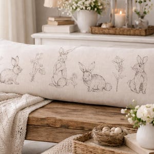 Könnte beinhalten: Ein langes, rechteckiges Kissen mit cremefarbenem Leinenbezug. Es zeigt Skizzen von Hasen und floralen Elementen in Grau. Das Kissen liegt auf einer Holzbank mit einer cremefarbenen Strickdecke. Das Kissen ist etwa 90 cm lang.
