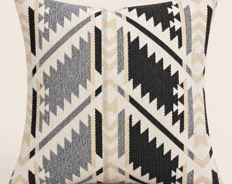 May include: A square throw pillow featuring a woven geometric pattern in black, charcoal gray, and beige. The design consists of repeating jagged, diamond-like shapes and vertical chevron stripes arranged in a symmetrical, repeating layout.
