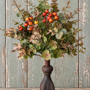 May include: A decorative topiary featuring a cluster of green and bronze eucalyptus leaves interspersed with small, round orange and red berries. The foliage is supported by a dark, weathered brown metal stand with a square base and carved details.