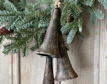 Op de afbeelding: Drie rustieke metalen bellen met een verweerde bronzen afwerking hangen aan een natuurlijk touw, geaccentueerd door een groene dennenkrans. De bellen hebben een conische vorm en hangen tegen een verweerde houten achtergrond.