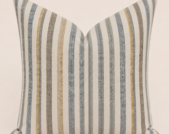 May include: A square throw pillow featuring a vertical striped pattern in shades of blue, tan, and grey. The fabric has a visible herringbone weave texture throughout, with alternating thin and thick stripes running across the entire surface.