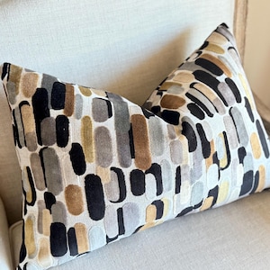 May include: A decorative cushion with a geometric pattern in shades of black, brown, grey, and gold. The cushion is made of a soft, plush fabric and has a subtle sheen.