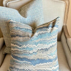 May include: Two decorative throw pillows with a blue and gray abstract pattern. The pillows are on a light beige armchair.