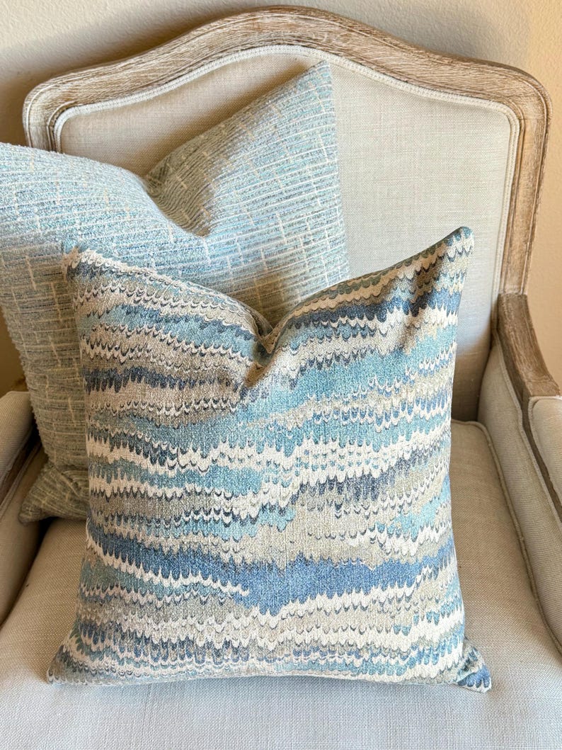 May include: Two decorative throw pillows with a blue and gray abstract pattern. The pillows are on a light beige armchair.