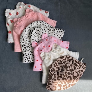 Op de afbeelding: Een verzameling baby zonnehoeden in verschillende patronen en kleuren. De hoeden omvatten een witte hoed met aardbeienprint, een roze hoed met een strik, een zwart-witte dalmatiërprint hoed, een roze hoed met rode hartjes en een bruine luipaardprint hoed.