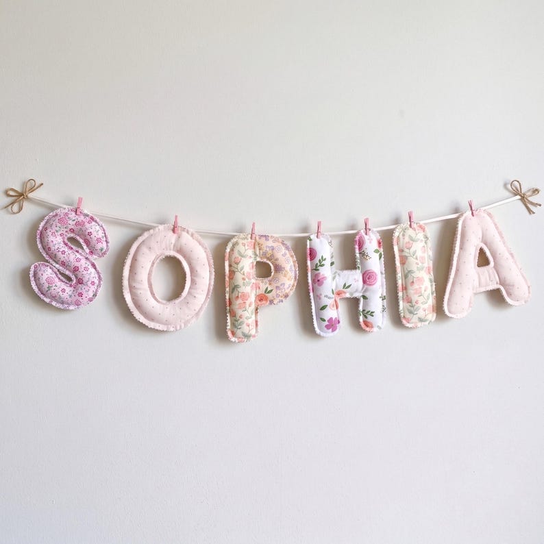 Puede incluir: Un cartel decorativo que deletrea el nombre SOPHIA con letras de tela acolchadas individuales. Cada letra presenta un patr&oacute;n floral o de puntos &uacute;nico en tonos rosa, melocot&oacute;n y blanco, suspendido de un cord&oacute;n blanco con clips.