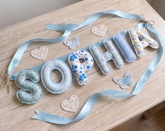 Puede incluir: Un adorno de pared con el nombre SOPHIA escrito en letras de tela acolchadas. Las letras presentan varios patrones florales, azules y blancos. El conjunto incluye corazones y mariposas de encaje blanco, unidos por una cinta de satén azul claro.