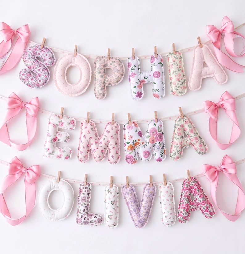 Puede incluir: Tres pancartas de tela horizontales con los nombres SOPHIA, EMMA y OLIVIA. Cada letra est&aacute; acolchada, estampada con diversos motivos florales en rosa, blanco y morado, y sujeta a una cinta de lunares con pinzas de madera y lazos rosas.