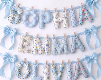 Puede incluir: Tres pancartas horizontales con letras de tela acolchada que forman los nombres SOPHIA, EMMA y OLIVIA. Cada letra está sujeta a una cinta de satén azul claro con pequeñas pinzas de madera. Los extremos de las cintas tienen lazos.