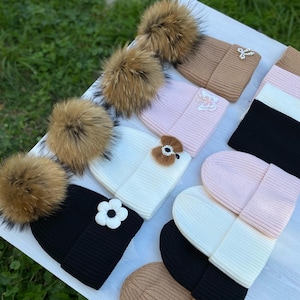 Könnte beinhalten: Eine Auswahl an gestrickten Wintermützen in verschiedenen Farben, darunter Schwarz, Weiß, Rosa und Beige. Einige Mützen haben große, flauschige braune Bommel, während andere mit floralen oder juwelenbesetzten Verzierungen versehen sind. Die Mützen sind auf einer weißen Oberfläche angeordnet.