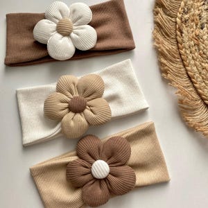 Könnte beinhalten: Drei gerippte Stirnbänder in Braun- und Cremetönen, jedes mit einer großen, plüschigen Blume verziert. Die Blumen haben ein geschichtetes Blütenblattdesign und einen kontrastierenden Mittelpunkt. Die Stirnbänder sind wahrscheinlich für Babys oder Kleinkinder.