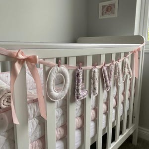 Puede incluir: Una guirnalda decorativa que deletrea el nombre OLIVIA con letras de tela individuales suspendidas de una cinta rosa claro. Cada letra presenta un dise&ntilde;o floral o estampado &uacute;nico en tonos rosa, morado y blanco, sujeto a la cinta con peque&ntilde;as pinzas de madera.