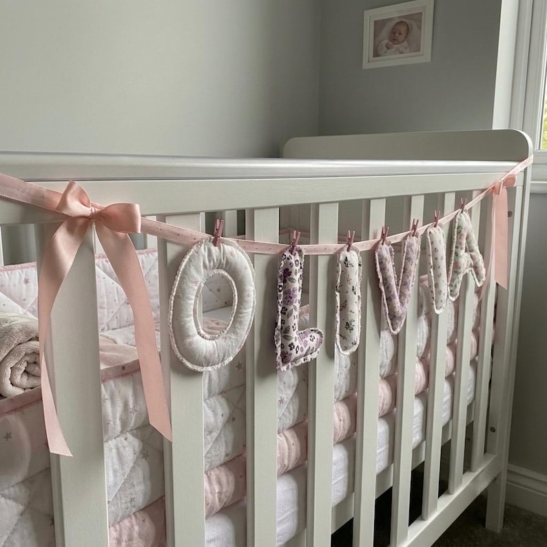 Puede incluir: Una guirnalda decorativa que deletrea el nombre OLIVIA con letras de tela individuales suspendidas de una cinta rosa claro. Cada letra presenta un dise&ntilde;o floral o estampado &uacute;nico en tonos rosa, morado y blanco, sujeto a la cinta con peque&ntilde;as pinzas de madera.