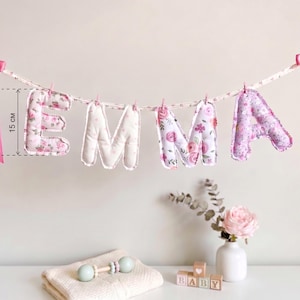 Puede incluir: Una guirnalda de tela que deletrea el nombre EMMA con letras acolchadas de estampado floral colgadas de una cinta. Cada letra mide 15 cm de altura. La guirnalda est&aacute; decorada con lazos de sat&eacute;n rosa en ambos extremos.