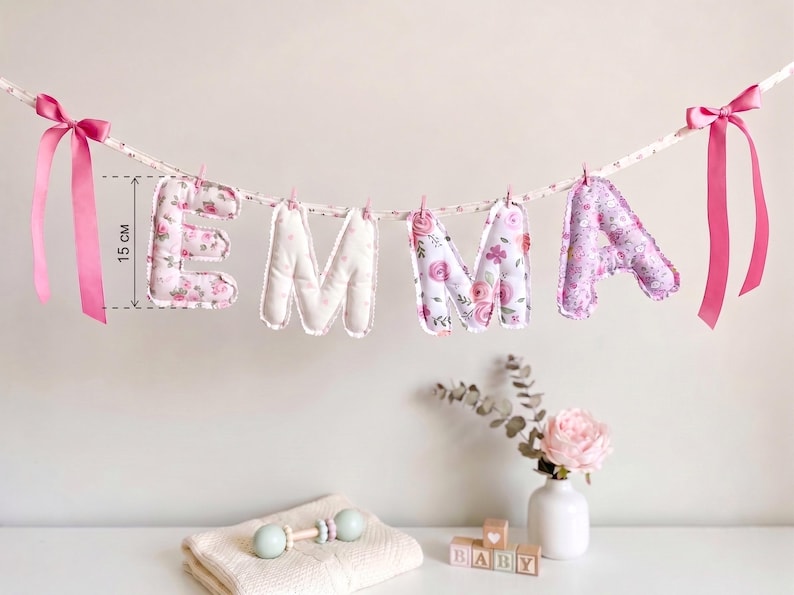Puede incluir: Una guirnalda de tela que deletrea el nombre EMMA con letras acolchadas de estampado floral colgadas de una cinta. Cada letra mide 15 cm de altura. La guirnalda est&aacute; decorada con lazos de sat&eacute;n rosa en ambos extremos.