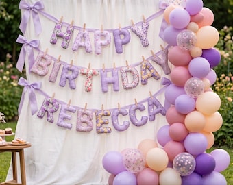 Puede incluir: Un telón de fondo de tela blanca con tres filas de letras de peluche colgantes que forman Happy Birthday Rebecca, decorado con cintas de color lavanda y un gran arco de globos en tonos morado, rosa y crema a la derecha.