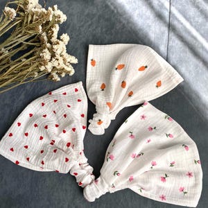 May include: Three white headbands with different patterns: red hearts, orange carrots, and pink flowers. The headbands are made of a soft, textured fabric and are knotted at the base. Dried flowers are in the background.