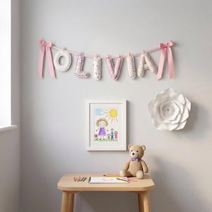 Puede incluir: Una guirnalda de tela con el nombre OLIVIA formado por letras acolchadas colgadas de una cinta rosa con lazos. Debajo hay un dibujo enmarcado de una familia, un oso de peluche beige con lazo morado y una mesa de madera con l&aacute;pices de colores.