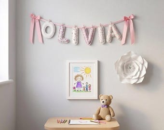 Puede incluir: Una guirnalda de tela que deletrea OLIVIA con letras acolchadas cuelga de una cinta rosa con lazos. Debajo, un dibujo enmarcado de una familia, un oso de peluche beige con pajarita morada y una mesa de madera con lápices de colores.