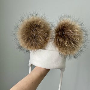 May include: A white winter hat with two large, fluffy, brown fur pom-poms. The hat has ear flaps and white ties. The pom-poms are a light brown color and are round in shape.