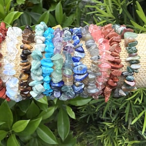 May include: A collection of gemstone chip bracelets in various colors, including red, white, brown, turquoise, blue, purple, pink, and green. The bracelets are displayed on a beige cylindrical surface, surrounded by green foliage.