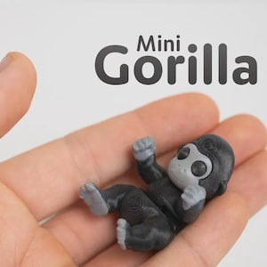 May include: A small, 3D-printed figurine of a gorilla with a black body and light grey face, hands, and feet. The figurine is positioned in a palm, and the text "Mini Gorilla" is printed in dark grey letters above the object.