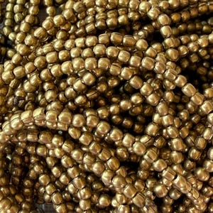 Puede incluir: Una gran colección de pequeñas cuentas redondas de color oro metálico dispuestas en múltiples hilos. Cada cuenta presenta una superficie lisa y reflectante con una costura sutil en el centro, creando una textura uniforme y densa en toda la imagen.