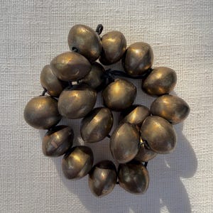 Puede incluir: Un conjunto de cuentas redondas de metal color bronce ensartadas en un cordón negro fino. Cada cuenta presenta una superficie lisa y ligeramente desgastada con sutiles variaciones de tono, creando una disposición circular y coherente.
