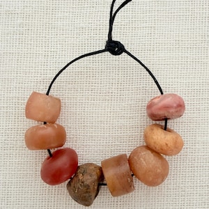 Peut inclure: Un arrangement circulaire de huit perles en pierre lisses et de formes irrégulières dans des tons pêche, orange, rouge et brun, enfilées sur un fin cordon noir noué au sommet, formant une boucle de perles minérales aux teintes naturelles.