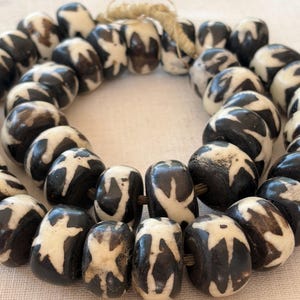 May include: A strand of round, dark brown beads featuring a white, star-like pattern etched into the surface. The beads are strung together on a light-coloured cord, creating a circular arrangement with visible texture and natural variations in the finish.