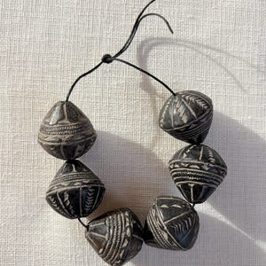 Puede incluir: Un collar de cinco cuentas cónicas de color gris oscuro ensartadas en un cordón negro fino. Cada cuenta presenta intrincados patrones geométricos tallados a mano, incluyendo líneas horizontales, triángulos y motivos de chevrón en la superficie.