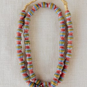 Puede incluir: Un collar de doble hebra con cuentas de vidrio en forma de barril con rayas horizontales en rojo, amarillo, verde y azul. Las cuentas están ensartadas en un cordón de fibra natural que forma un lazo en la parte superior, creando un diseño circular.