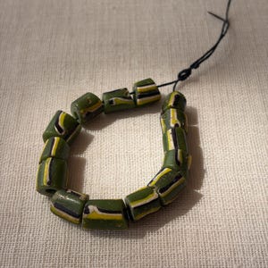 Può includere: Una collana composta da undici perline cilindriche infilate su un sottile cordino nero. Ogni perla presenta una base color verde oliva con strisce orizzontali bianche, gialle e nere. Le perle hanno un aspetto leggermente irregolare.