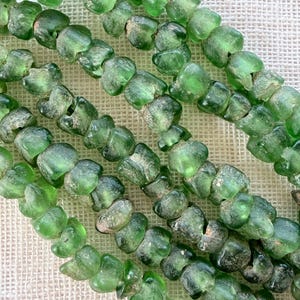 Può includere: Una fila di perline in vetro verde smeraldo traslucido con forme irregolari e arrotondate e superfici usurate. Le perline sono infilate insieme in una linea continua, mostrando variazioni di chiarezza e consistenza su ogni pezzo.