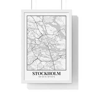 May include: A framed print featuring a detailed black and white map of Stockholm, Sweden. The city map is encased in a white frame and includes the city name and coordinates at the bottom. The print is suspended by a white cord.