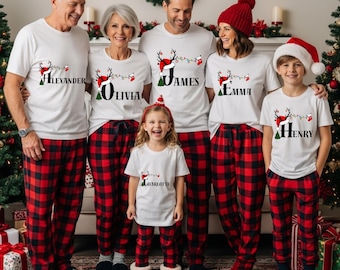 May include: A family wearing matching Christmas pajamas. The white t-shirts have a reindeer design with names printed on them. The red and black plaid pajama pants complete the festive look. A Christmas tree and decorations are in the background.