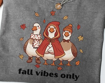 May include: A charcoal gray t-shirt featuring a graphic of three geese wearing autumn accessories, including a scarf, a red hooded cloak, and a beanie. The text "fall vibes only" is printed below the geese, surrounded by falling maple leaves.