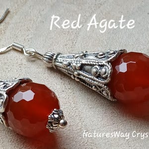 May include: Earrings featuring red agate gemstones with a faceted design. The earrings have silver-coloured, decorative metalwork. The image includes the text "Red Agate" and "NaturesWay Crystals".