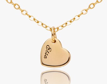 May include: A gold-colored necklace with a heart-shaped pendant. The pendant has the name "Elsa" engraved on it. The necklace chain is delicate and the pendant reflects on the surface below.