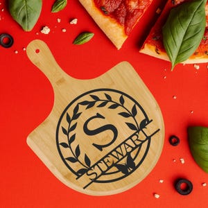 Może przedstawiać: Bambusowa deska do serwowania pizzy z uchwytem. Deska posiada czarny okrągły wzór z literą "S" i nazwiskiem "STEWART" z motylem. Kawałki pizzy, liście bazylii i oliwki otaczają deskę na czerwonej powierzchni.