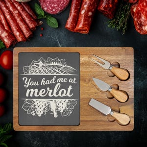 May include: Wooden charcuterie board with a dark gray slate insert that reads "You had me at merlot." Includes three cheese knives and a fork. Salami and tomatoes are arranged around the board.