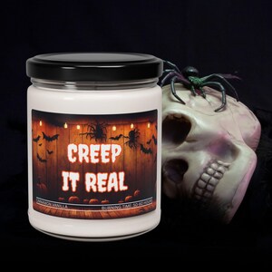 Può includere: Una candela in barattolo di vetro bianco con coperchio nero, etichettata "CREEP IT REAL" a tema Halloween. L'etichetta presenta pipistrelli, ragni e zucche su uno sfondo di legno. Un teschio decorativo con un ragno è accanto alla candela.