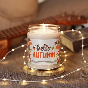 Pode incluir: Uma vela acesa em um frasco de vidro com um rótulo que diz "hello AUTUMN" em marrom e laranja. A vela está em uma base redonda de madeira, cercada por luzes de corda. A vela tem um aroma de baunilha e canela.