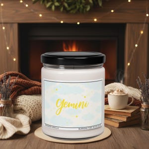 May include: A white candle in a glass jar with a black lid. The label features the word "Gemini" in yellow script, with a light blue cloud and star design. The candle is on a wooden coaster, with a cozy fireplace in the background.
