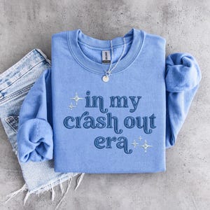 Könnte beinhalten: Hellblaues Sweatshirt mit dem Text "in my crash out era" in dunkelblauer, stilisierter Schrift. Das Sweatshirt ist gefaltet, die Ärmel sind zu sehen, und eine silberne Halskette mit Anhänger liegt obenauf. Eine Jeans ist daneben.