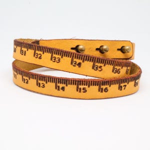 May include: A tan leather wrap bracelet with a ruler design. The bracelet is marked with inch measurements and has three brass-coloured studs. The bracelet is wrapped around itself.